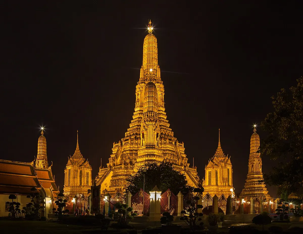 Wat Arun in Bangkok illuminated at night, framed by trees.