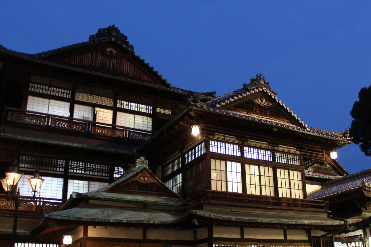 青い夕暮れの空の下、木造の道後温泉本館の窓に温かな明かりがともっています。