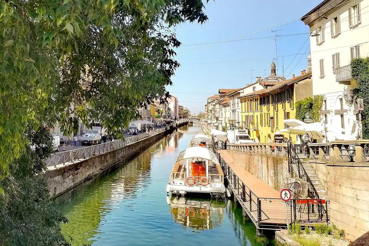 The Naviglio Grande on a clear day, seen past tree branches, with a canal boat moored at a pier and yellow houses along the water.
