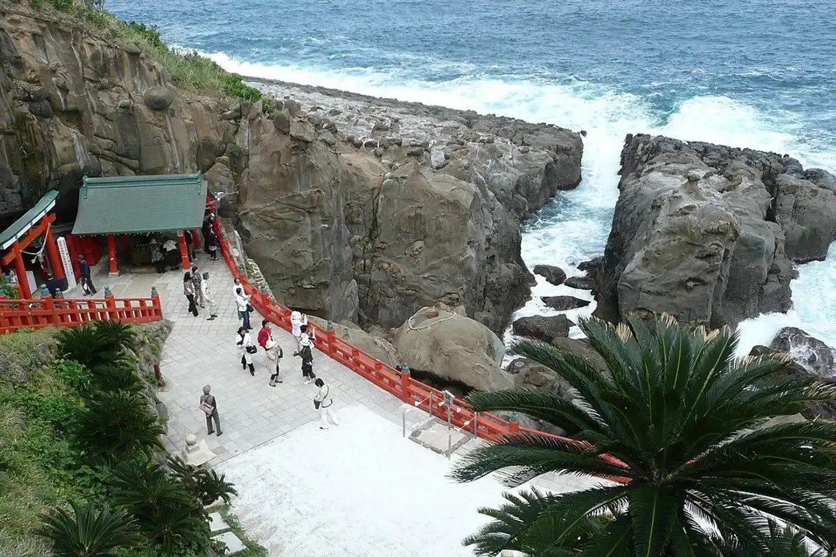 Red railings and shrine buildings at the mouth of a sea cave, with waves breaking on the rocks beside them.