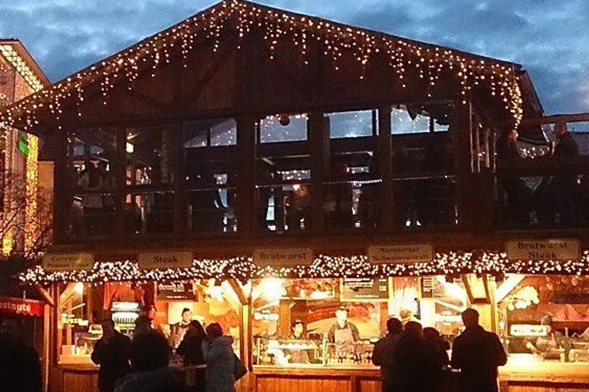 A wooden market stall strung with lights at dusk, with silhouettes of people in front.