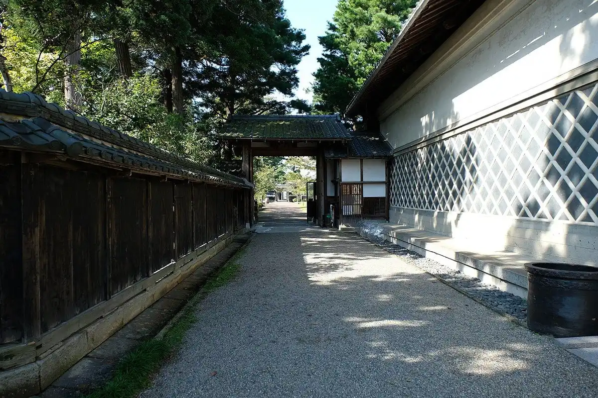 瓦を載せた黒い木の塀と、ひし形模様の白い蔵の壁の間を門まで続く砂利道（北方文化博物館）。
