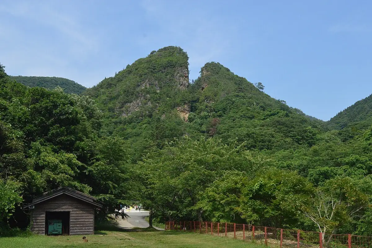 緑の森の上で山頂がV字に割れた佐渡金山の道遊の割戸と、手前の芝生の小さな木の小屋。