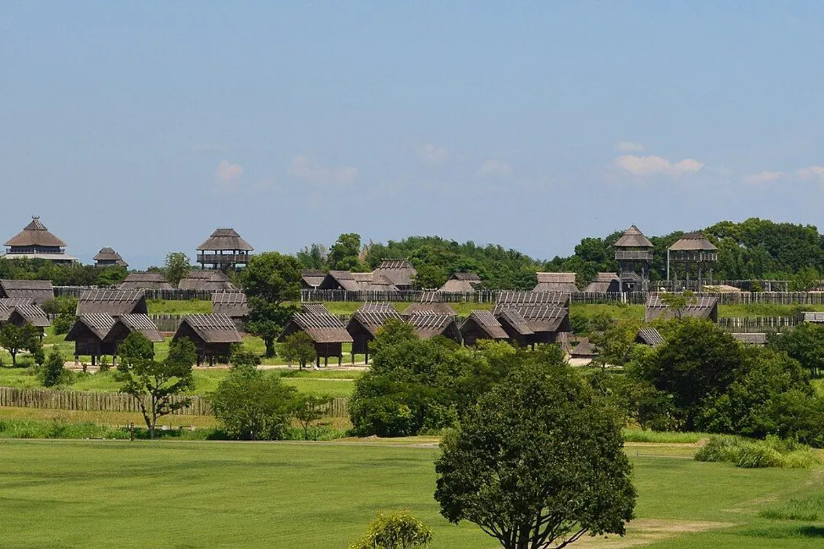 Reconstructed thatched buildings and lookout towers of the Yoshinogari moated settlement beyond a wide lawn.