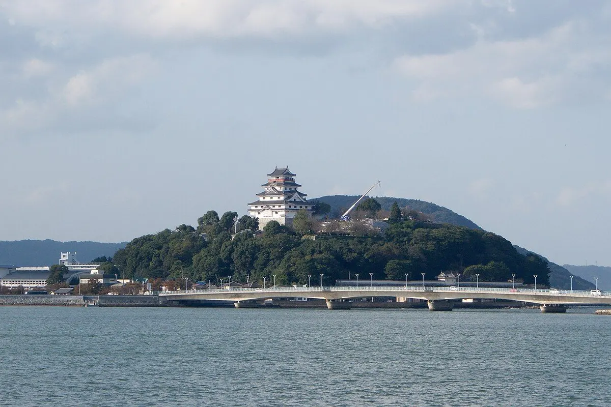 The white keep of Karatsu Castle on a wooded hill across the water, with a long bridge.