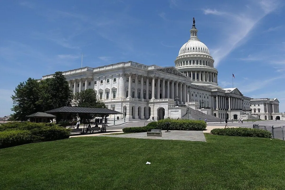 The white dome and Senate wing of the U.S. Capitol seen across a green summer lawn under a blue sky.