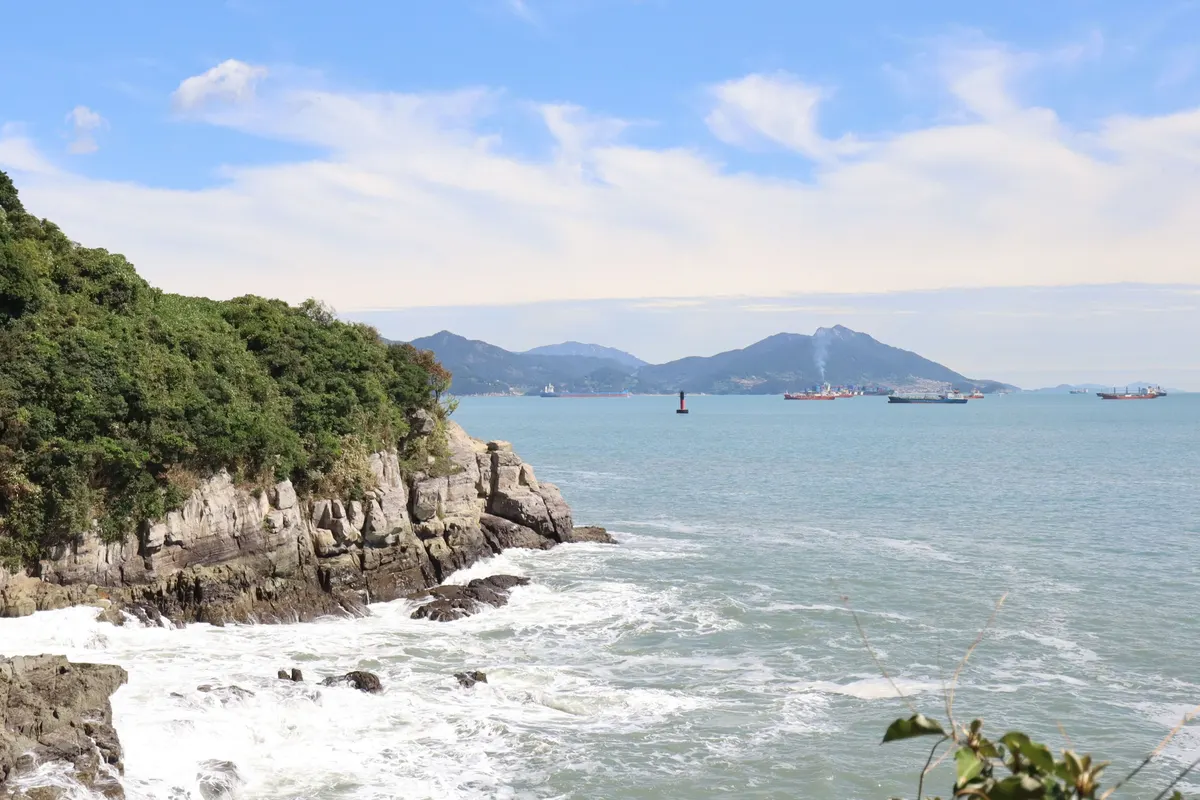 森に覆われた梧桐島の岩の海岸に波が砕け、沖には島々と船が見えます。