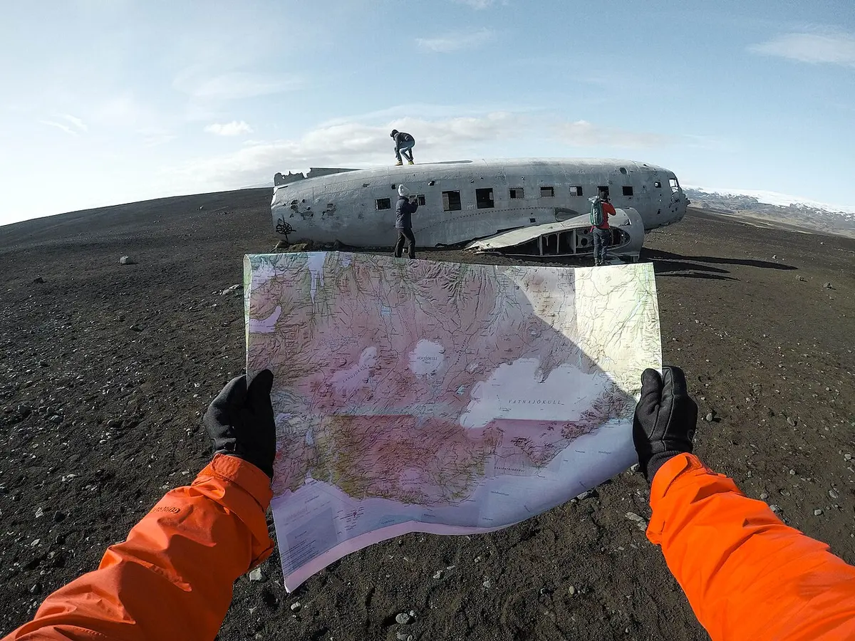 黒い砂の上の飛行機の残骸の前で、手袋をした手で紙の地図を広げている様子。
